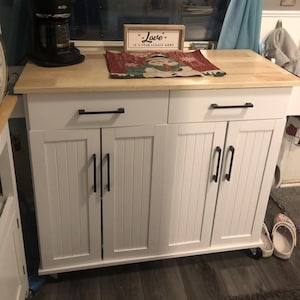 May include: A white kitchen island with a light wood countertop. It has black handles and a cabinet door design. A coffee maker and a decorative sign that reads "Love is a four legged word" are on top. A Christmas-themed mat is also on the countertop.