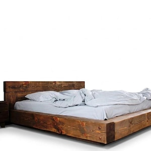 May include: A rustic wooden bed frame with a headboard and a matching nightstand. The bed is made of dark-stained wood and has a low profile. The bedding includes a white comforter and a patterned pillow. The nightstand has two drawers.