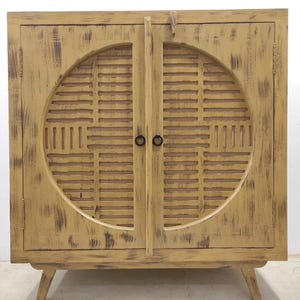 Puede incluir: Un aparador de dos puertas desgastado en un tono amarillo apagado. El mueble presenta un gran diseño circular en las puertas con un patrón de listones. Tiene pequeños tiradores circulares negros y se apoya en cuatro patas anguladas.