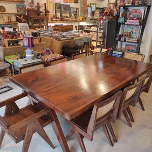 Puede incluir: Una gran mesa de comedor rectangular de madera con un acabado marrón oscuro. La mesa está rodeada de sillas de madera a juego con respaldos de mimbre tejido. La escena tiene lugar en una tienda bien iluminada.