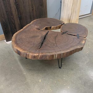 Puede incluir: Una mesa de centro única con borde natural. La mesa está hecha de madera marrón oscuro con una forma natural y orgánica. Cuenta con una gran superficie irregular sostenida por patas de horquilla negras. La veta de la madera y las grietas añaden encanto rústico.
