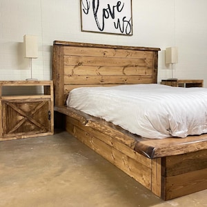 Op de afbeelding: Een rustiek houten bedframe met een bijpassend hoofdbord en nachtkastje. Het bed heeft een natuurlijke houten afwerking en een laag profiel. Het nachtkastje heeft een enkele deur met een X-vormig ontwerp. Een wit dekbed bedekt het bed.