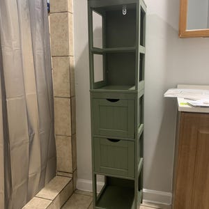 May include: A tall, olive green bathroom storage cabinet with open shelves and two drawers. The cabinet has a rectangular shape and is made of wood. The drawers have a recessed panel design and small, round pulls. The cabinet is free-standing.
