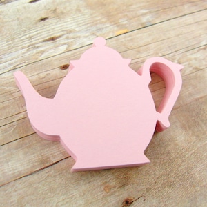 May include: A pink paper teapot cutout, layered to create a 3D effect. The teapot is sitting on a light brown wooden surface.