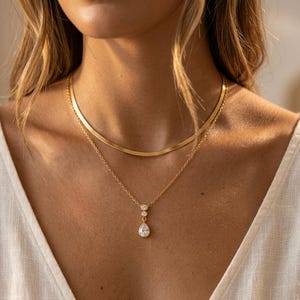 Könnte beinhalten: Ein goldfarbenes Halsketten-Set, bestehend aus einer Fischgrätenkette und einer Anhängerkette mit drei klaren, runden und tropfenförmigen Steinen. Der Schmuck wird über einem weißen Oberteil getragen.