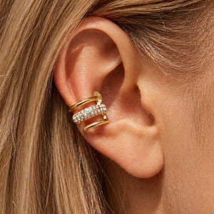 May include: Gold-toned ear cuff with a triple-band design. The cuff features a row of sparkling clear crystals in the center band, adding a touch of glamour. The jewelry is worn on the ear, showcasing its elegant style.