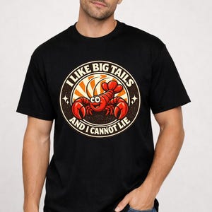 Op de afbeelding: Zwart T-shirt met een cirkelvormige afbeelding van een cartoon kreeft en de tekst "I LIKE BIG TAILS AND I CANNOT LIE". De kreeft is rood met een lachend gezicht, tegen een zonneschijnontwerp.