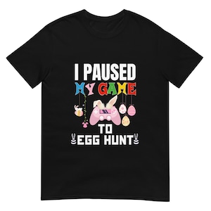 Op de afbeelding: Zwart t-shirt met de witte tekst "I PAUSED MY GAME TO EGG HUNT". Het ontwerp bevat een roze gamecontroller met konijnenoren, paaseieren en andere paasgerelateerde afbeeldingen in verschillende kleuren.