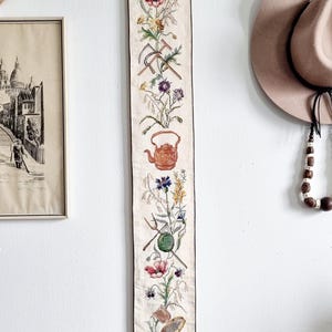 May include: A white fabric wall hanging with a brown trim and fringe. The hanging features a vertical embroidered design of flowers, a teapot, a cart, and gardening tools.