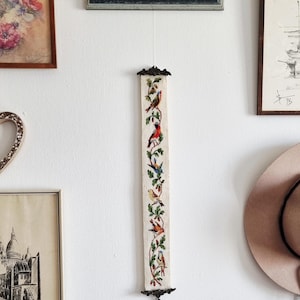 Könnte beinhalten: Ein langer, schmaler, bestickter Wandbehang mit bunten Vögeln auf belaubten Zweigen. Das Kunstwerk wird von dekorativen Metallbeschlägen oben und unten gerahmt. Der Hintergrund ist cremefarben.