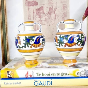 Może przedstawiać: Dwa dekoracyjne wazony ceramiczne z uchwytami. Wazony są białe z żółtymi, niebieskimi, zielonymi i pomarańczowymi wzorami kwiatowymi. Stoją na książkach z kolorowymi okładkami. W tle widoczna jest oprawiona grafika.