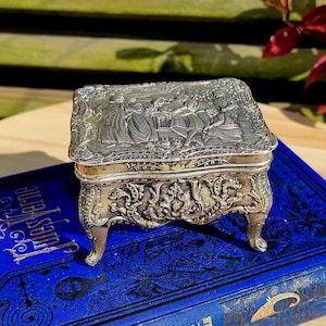 Puede incluir: Una caja de alhajas de plata con un diseño ornamentado y tres patas. La caja está decorada con una imagen en relieve de personas en una escena. La caja está colocada sobre un libro azul.