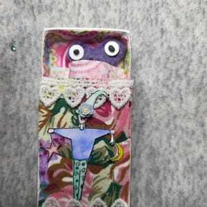 May include: A whimsical matchbox art piece featuring a miniature figure with a blue shirt and patterned pants. The figure is adorned with a star and crescent moon. The matchbox interior has a pink floral design, and the top has a purple eye mask.