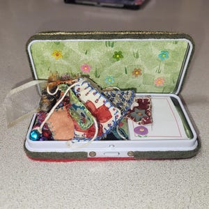 Può includere: Scatola di metallo aperta con interno verde a motivi floreali e base in feltro rosso. Contiene ritagli di tessuto colorati, perline e una piccola carta. Un nastro bianco è attaccato.