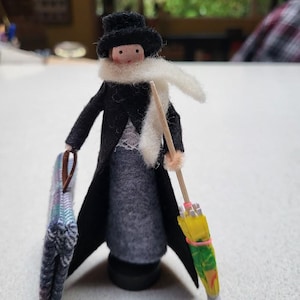 Pode incluir: Boneca artesanal vestida com casaco preto, chapéu e cachecol branco, segurando uma bolsa estampada e um guarda-chuva em miniatura. A boneca tem cabeça de madeira e saia cinza. O guarda-chuva é amarelo e verde.
