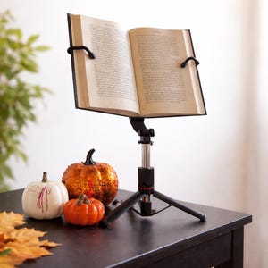 May include: A black adjustable book stand with a tripod base holding an open hardcover book. The stand features a central silver telescopic pole and two black flexible arms that secure the pages of the book in an open, readable position.