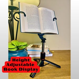 May include: A black and silver height-adjustable book display holding an open book. The display has a tripod base and curved arms to hold the book open. The text "Height Adjustable Book Display" is in a red banner.