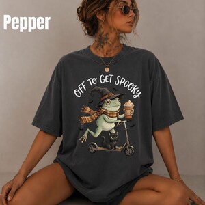 May include: A charcoal gray t-shirt featuring a graphic of a green frog wearing a witch hat and a plaid scarf, riding a scooter with a black cat. The frog holds a coffee cup, and the text reads OFF TO GET SPOOKY. The shirt color is labeled Pepper.