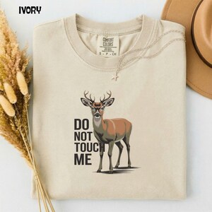 Könnte beinhalten: Elfenbeinfarbenes T-Shirt mit einer Hirschillustration und dem Text "DO NOT TOUCH ME". Der Hirsch ist braun und beige mit Geweih. Eine goldene Halskette mit Sternanhänger befindet sich auf dem T-Shirt.