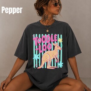 May include: A charcoal gray short-sleeve t-shirt featuring a graphic print of a light orange kangaroo. The design includes the text "TOODLE-LOO" in pink letters, set against a background of vertical teal stripes and light blue and yellow stars.