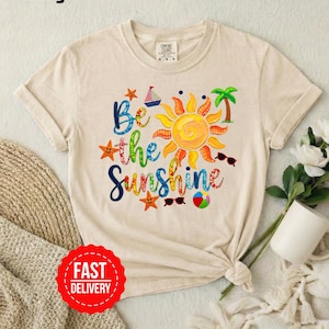 May include: Ivory t-shirt featuring the phrase "Be the Sunshine" in vibrant colours. The design includes a sun, sailboat, palm tree, sunglasses, starfish, and a beach ball. The shirt has a round neck and short sleeves.