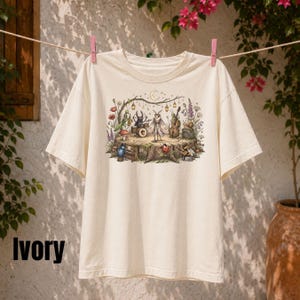 Op de afbeelding: Een ivoorkleurig t-shirt met korte mouwen en een gedetailleerde grafische print van diverse insecten die muziekinstrumenten bespelen op een boomstronk. De illustratie bevat een mot, kever en slak omringd door bloemen, hangende lantaarns en een maan.