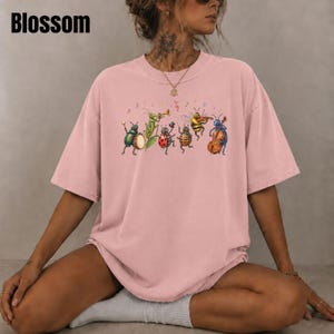 May include: A dusty pink short-sleeve t-shirt featuring a graphic print of various insects playing musical instruments, including a beetle with a drum, a grasshopper with a trumpet, a ladybug, a bee with a violin, and a blue insect with a cello.