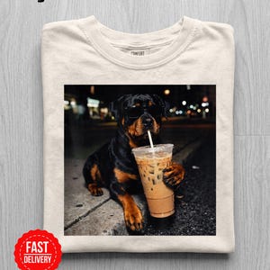 Könnte beinhalten: Ein elfenbeinfarbenes T-Shirt mit einem Grafikdruck eines schwarz-braunen Rottweilers, der eine schwarze Sonnenbrille trägt. Der Hund sitzt nachts auf einem Bordstein und hält einen durchsichtigen Plastikbecher mit Eiskaffee und einem weißen Strohhalm.