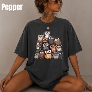 Puede incluir: Una camiseta de manga corta gris carb&oacute;n con un gran estampado gr&aacute;fico de un grupo de varios b&uacute;hos en tonos marr&oacute;n, gris y blanco. La palabra Pepper est&aacute; impresa en blanco en la esquina superior izquierda de la imagen. Cuello redondo.