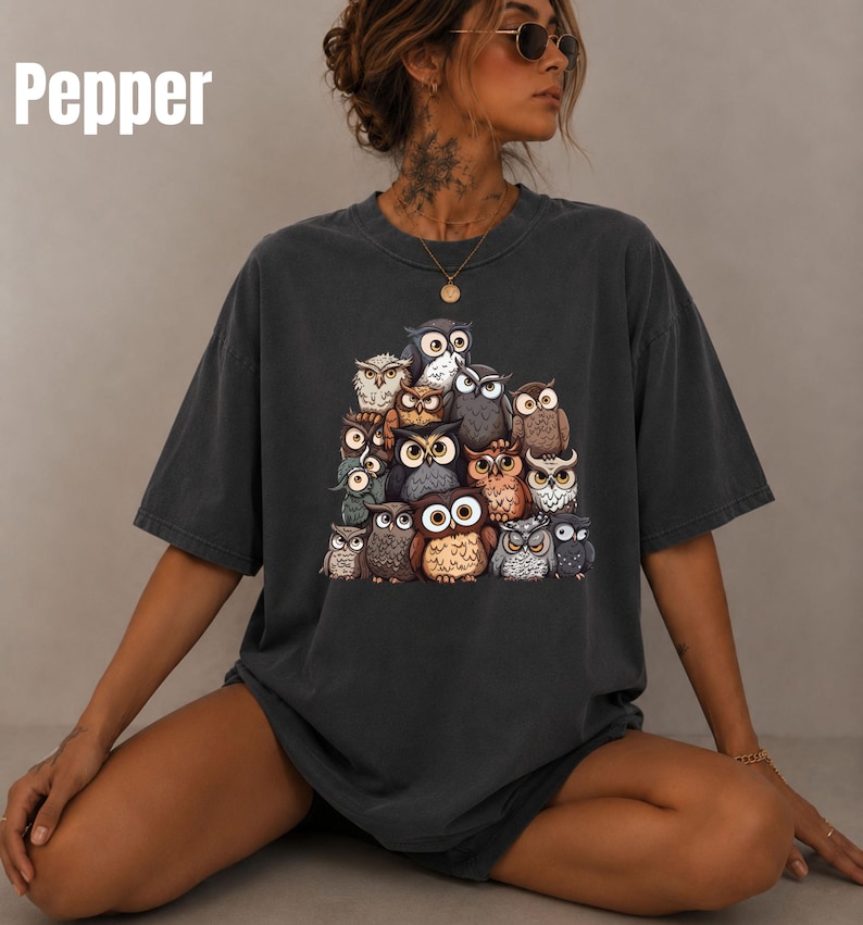 Puede incluir: Una camiseta de manga corta gris carb&oacute;n con un gran estampado gr&aacute;fico de un grupo de varios b&uacute;hos en tonos marr&oacute;n, gris y blanco. La palabra Pepper est&aacute; impresa en blanco en la esquina superior izquierda de la imagen. Cuello redondo.