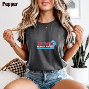 May include: Dark gray t-shirt with the text "BIG FAN OF HUMAN RIGHTS" in red and blue. The shirt has short sleeves and a crew neck. The word "Pepper" is in the top left corner.