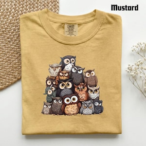 Puede incluir: Una camiseta de color amarillo mostaza con un estampado gr&aacute;fico de un gran grupo de b&uacute;hos variados en tonos marrones, grises y blancos. La camiseta tiene un cuello redondo y una etiqueta de la marca Comfort Colors en el interior.