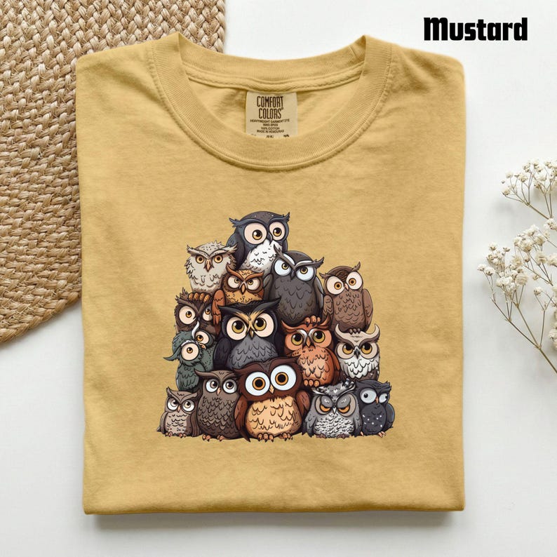 Puede incluir: Una camiseta de color amarillo mostaza con un estampado gr&aacute;fico de un gran grupo de b&uacute;hos variados en tonos marrones, grises y blancos. La camiseta tiene un cuello redondo y una etiqueta de la marca Comfort Colors en el interior.