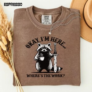 Puede incluir: Camiseta color espresso con un mapache sosteniendo una taza de café y una espada. El texto en la camiseta dice: "Okay, I'm here... Where's the work?" Un collar plateado con un colgante de estrella está sobre la camiseta.