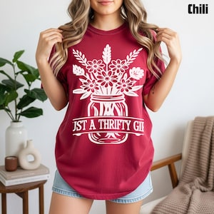 Könnte beinhalten: Ein weinrotes T-Shirt mit einem weißen Blumendesign in einem Einmachglas und dem Text "Just a Thrifty Girl". Das Shirt ist ein lässiger Stil, ideal für den Alltag. Das Shirt ist einfarbig.