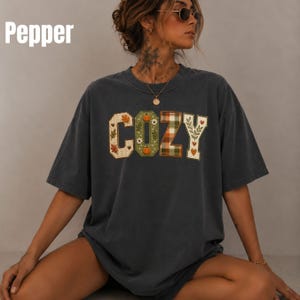 Pode incluir: Uma t-shirt de manga curta em cinzento-carvão com a palavra COZY impressa em letras grandes de bloco no peito. Cada letra contém um padrão sazonal único, incluindo folhas de outono, abóboras, xadrez verde e desenhos botânicos florais.