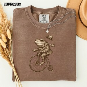 Könnte beinhalten: Espressofarbenes T-Shirt mit einer skurrilen Illustration eines Frosches auf einem Hochrad, der einen Schal und eine Jacke trägt. Das Shirt hat ein Comfort Colors-Etikett und eine silberne Halskette mit einem Sternanhänger. Das Wort "ESPRESSO" ist links oben aufgedruckt.