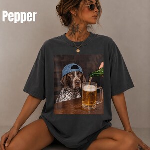 May include: A charcoal gray t-shirt featuring a graphic print of a brown and white spotted dog wearing a blue baseball cap. The dog sits at a wooden table while a green bottle pours beer into a glass mug filled with golden liquid and foam.