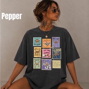 May include: A charcoal gray short-sleeve t-shirt featuring a grid of nine colorful postage stamp-style graphics. The designs include various cartoon characters and text such as 100% MEAT, ASTROPHAGE, JAZZ HANDS, AMAZE, FIT MY BUMP, WHY STUPID QUESTION, YOU SLEEP I WATCH.