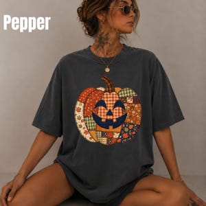 May include: A charcoal grey short-sleeve t-shirt featuring a large, central graphic of a jack-o'-lantern pumpkin composed of various colourful patchwork patterns, including plaid, gingham, and autumn leaf prints. The text Pepper is visible.