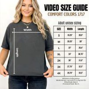 May include: A dark gray Comfort Colors 1717 unisex t-shirt with a size guide chart. The chart shows measurements for sizes S through 4X, including width and length in inches. The shirt is worn by a person.