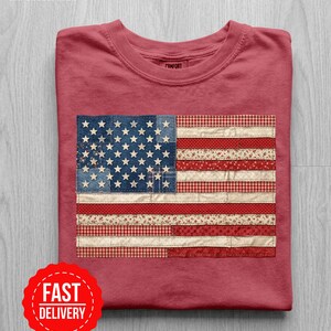 Puede incluir: Camiseta de color carmesí con un diseño de bandera americana de patchwork. La bandera incorpora patrones de mezclilla, florales y cuadros. La camiseta tiene la palabra "Comfort" impresa en el cuello. Una pegatina roja de "Fast Delivery" está en la esquina inferior izquierda.
