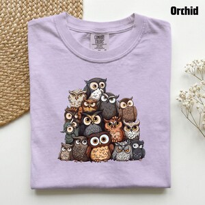 Puede incluir: Una camiseta de color morado claro con un estampado gr&aacute;fico de un gran grupo de b&uacute;hos en varios tonos de marr&oacute;n, gris y blanco. La camiseta tiene un cuello redondo y una etiqueta de la marca Comfort Colors en el interior del cuello.