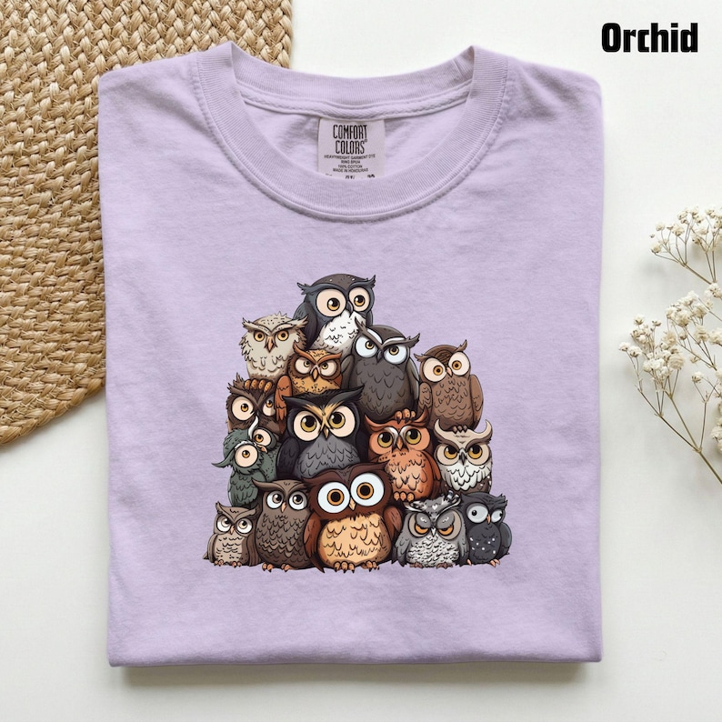 Puede incluir: Una camiseta de color morado claro con un estampado gr&aacute;fico de un gran grupo de b&uacute;hos en varios tonos de marr&oacute;n, gris y blanco. La camiseta tiene un cuello redondo y una etiqueta de la marca Comfort Colors en el interior del cuello.