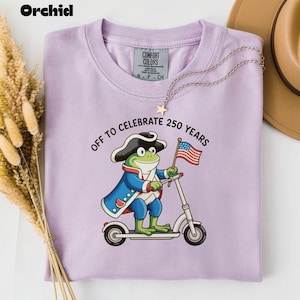 Puede incluir: Una camiseta lila Comfort Colors con un gráfico de una rana con sombrero tricornio montada en un scooter y sosteniendo una bandera estadounidense. El texto dice "OFF TO CELEBRATE 250 YEARS". Un collar dorado con un colgante de estrella está sobre la camiseta.