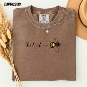 以下が含まれることがあります： 「Let it bee」のフレーズと蜂のグラフィックが特徴の、エスプレッソカラーのComfort Colors Tシャツ。星のペンダントが付いたシルバーのネックレスがシャツにかけられています。麦わら帽子が背景に一部見えています。