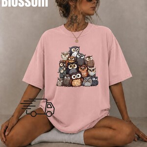Puede incluir: Una camiseta de manga corta en color rosa empolvado con un estampado gr&aacute;fico central de un grupo de varios b&uacute;hos en tonos marrones, grises y blancos. La palabra Blossom est&aacute; impresa en blanco en la esquina superior izquierda.