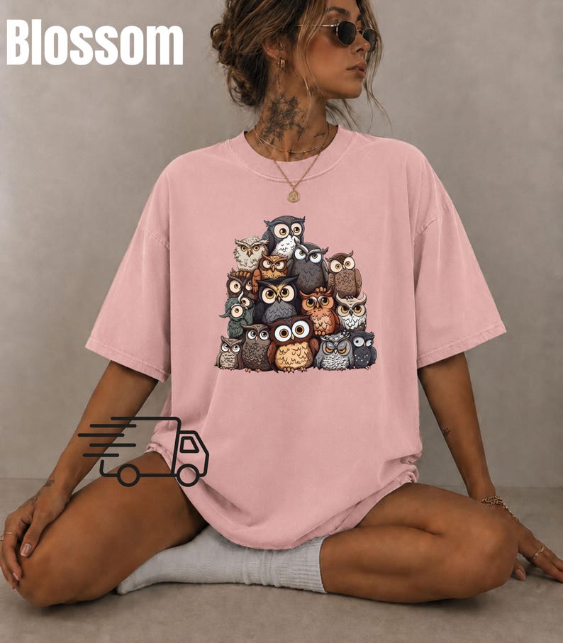 Puede incluir: Una camiseta de manga corta en color rosa empolvado con un estampado gr&aacute;fico central de un grupo de varios b&uacute;hos en tonos marrones, grises y blancos. La palabra Blossom est&aacute; impresa en blanco en la esquina superior izquierda.