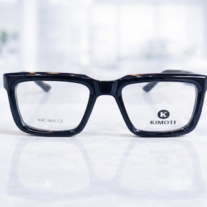 Puede incluir: Gafas negras y carey con montura cuadrada. Las gafas tienen lentes transparentes y el texto "KAC 0611 C2" está impreso en la patilla izquierda. La marca "KIMOTI" está impresa en la lente derecha.