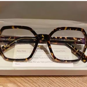 Könnte beinhalten: Brille mit Schildpattmuster und geometrischem Rahmen in einem offenen, weißen Etui. Die Brille hat klare Gläser und ein dunkelbraunes und beiges Muster. Eine weiße Karte mit Text ist darunter sichtbar.